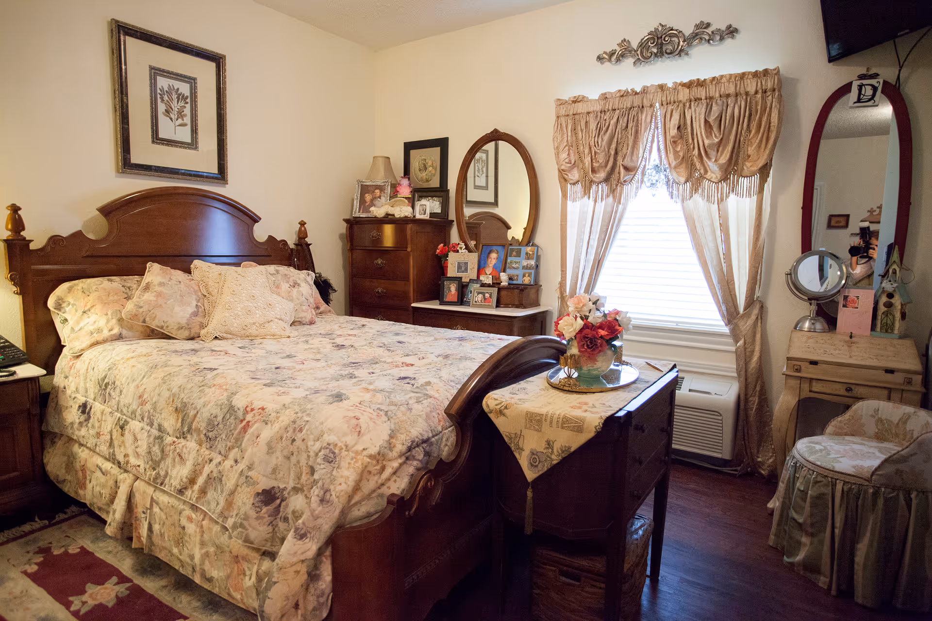 A cozy bedroom with a wooden bed frame and floral bedding. There is a wooden dresser with framed photos and a lamp, an oval mirror on a vanity table, a window with beige curtains, and a small cushioned chair. The room has warm lighting and wooden flooring.