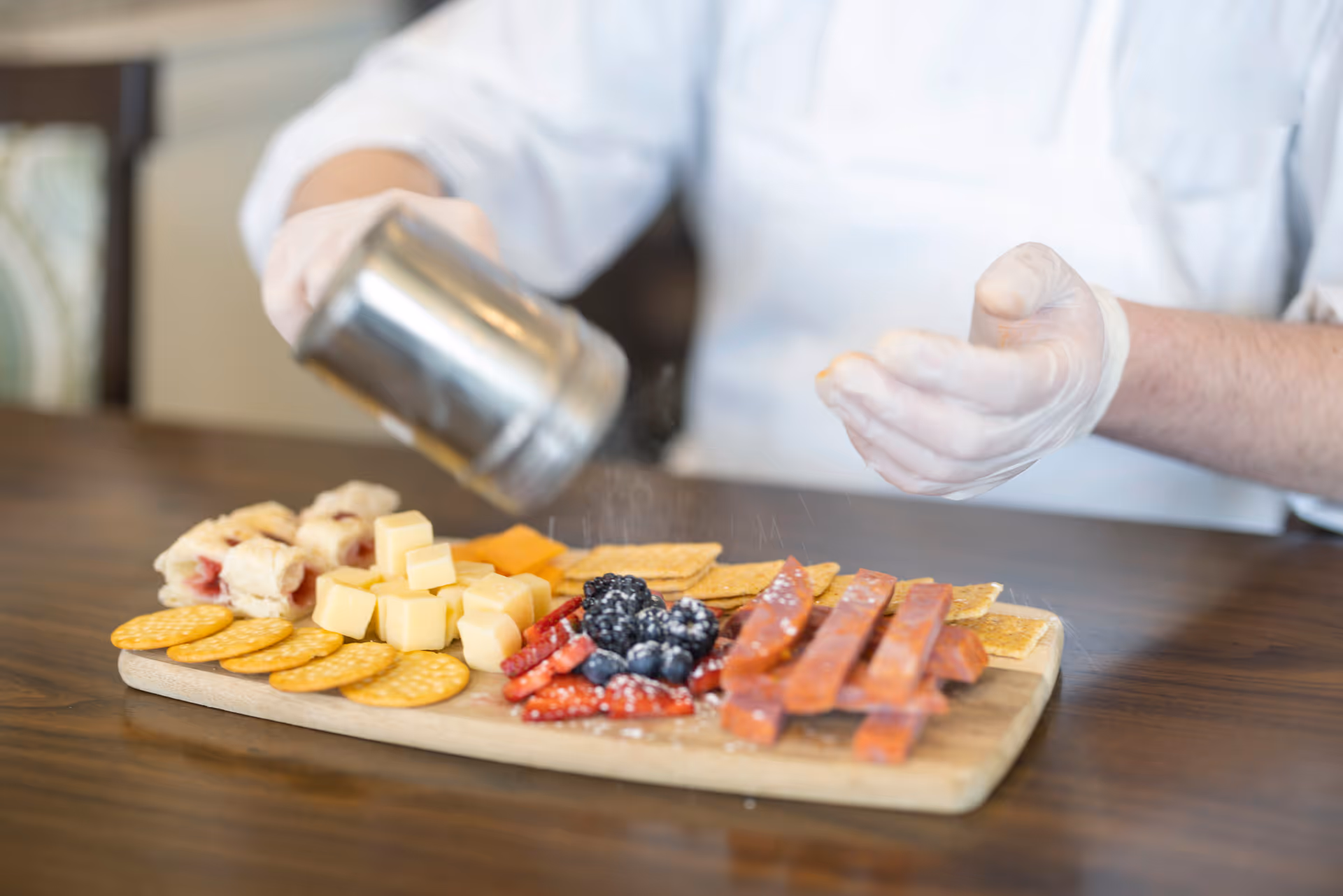 A person wearing gloves is sprinkling seasoning over a wooden board with an assortment of snacks including crackers, cheese cubes, berries, and meat sticks on a wooden table.