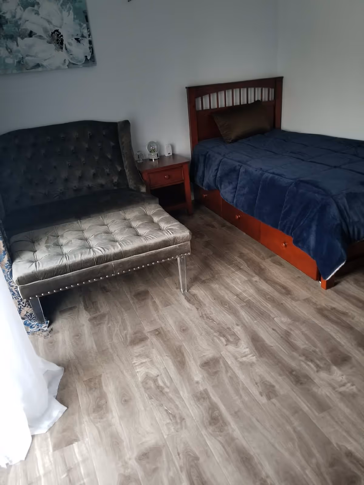 A bedroom with a wooden bed frame and a dark blue comforter, a brown pillow, a wooden nightstand with a glass dome decoration and a small device, a gray tufted loveseat with clear legs, and light wood flooring.
