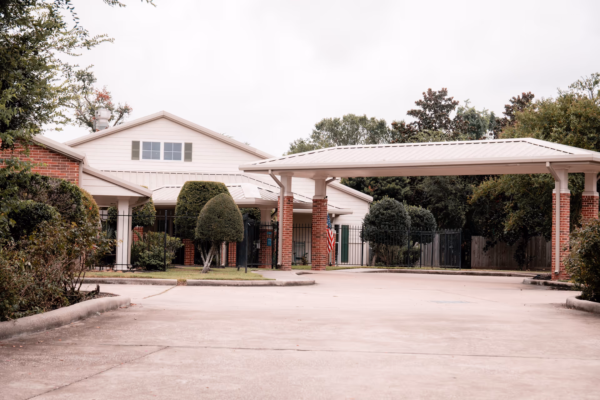 Front entrance of a light-colored building with a covered porte-cochere, driveway, and trimmed landscaping.
