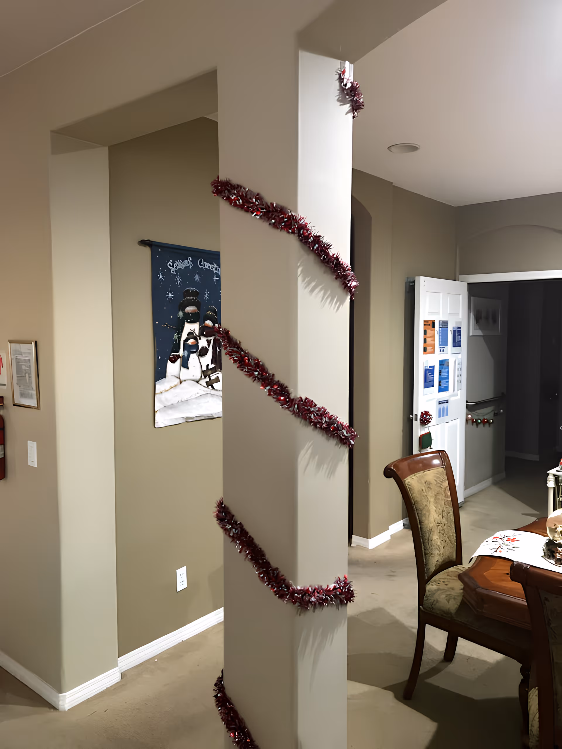 Interior view of a dining area in a senior living facility with a decorated pillar wrapped in red and silver garland. There is a wooden dining table with upholstered chairs, a festive table runner, and a snowman-themed wall hanging in the background. The walls are painted beige, and there is a white door with notices posted on it.