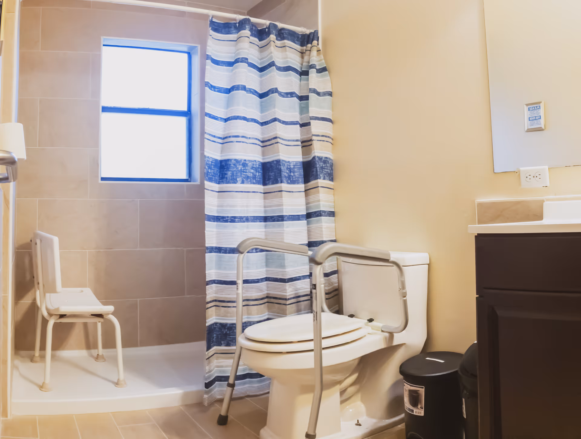 Bathroom with striped shower curtain, toilet with grab bars, shower chair, and sink cabinet.