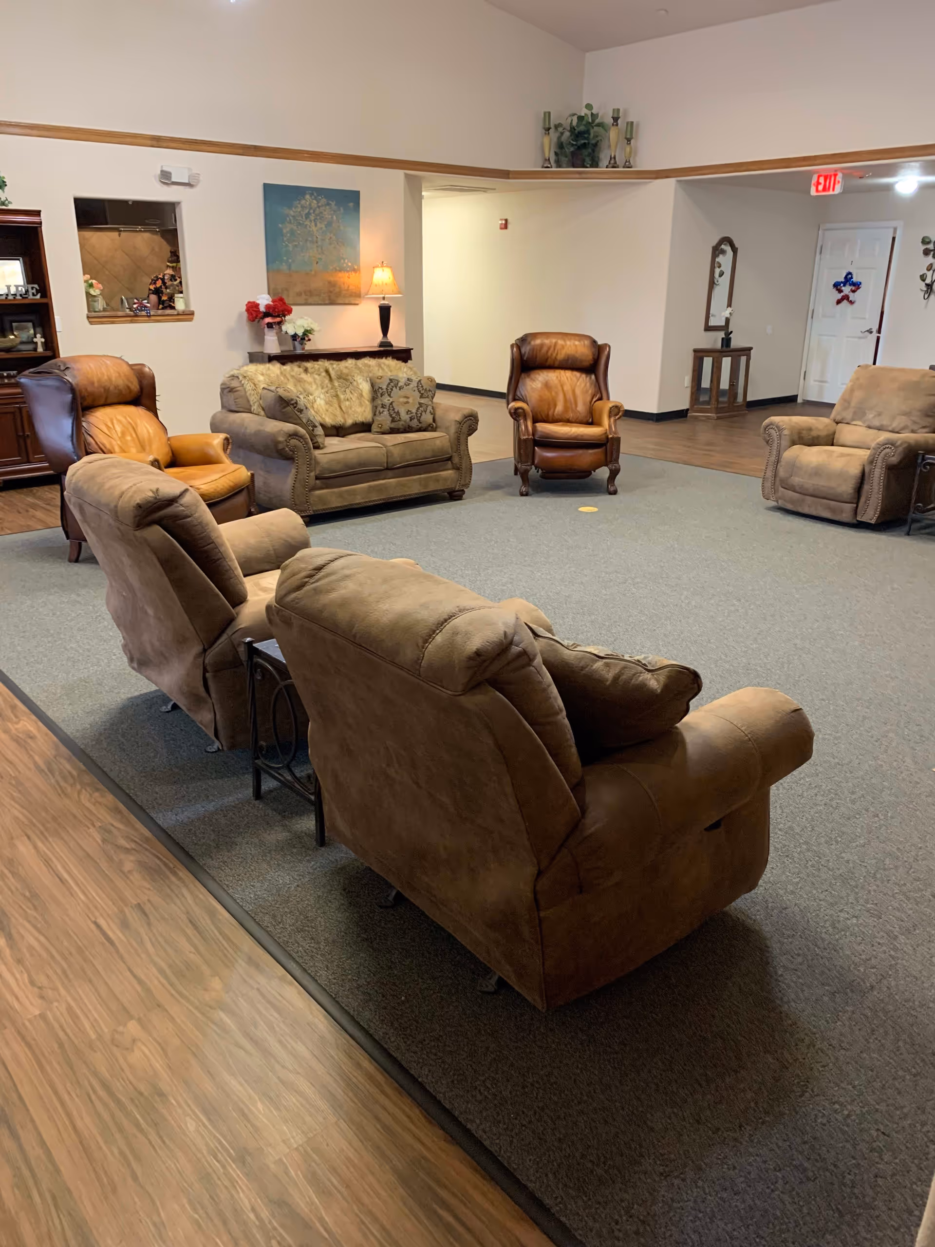 A senior living facility common area with several comfortable armchairs and a loveseat arranged in a circle on a carpeted floor. The room has wood flooring around the carpet, a side table with a lamp, wall art, and decorative plants on a shelf above the entrance. There is a small window opening to another room and a door decorated with a star-shaped ornament in the background.