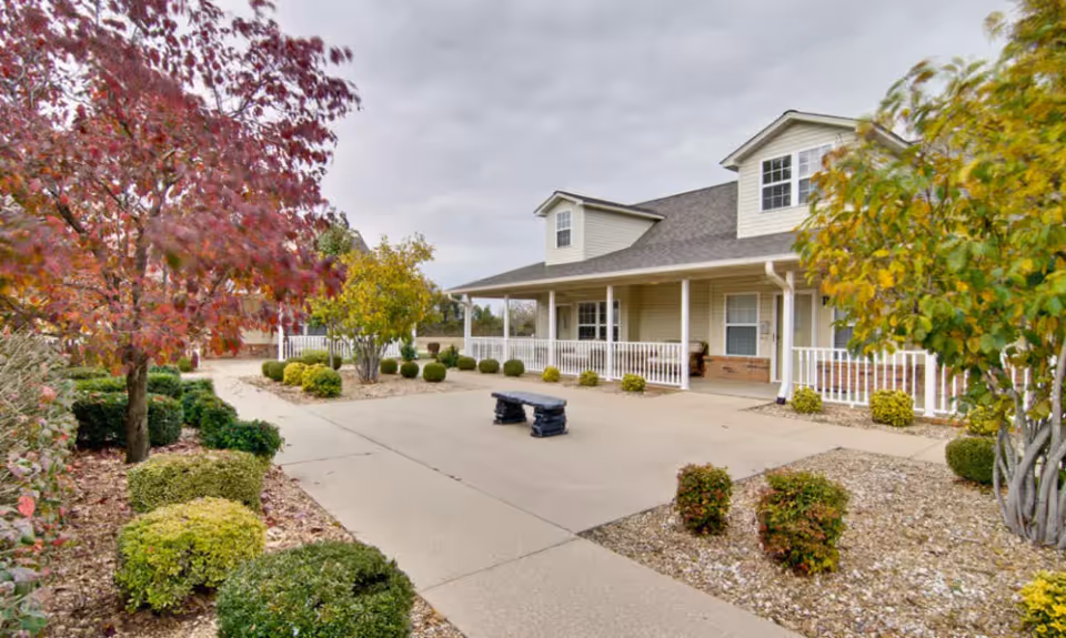 Front exterior of a senior living building with a covered porch, paved entrance, bench, and landscaped shrubs and trees.