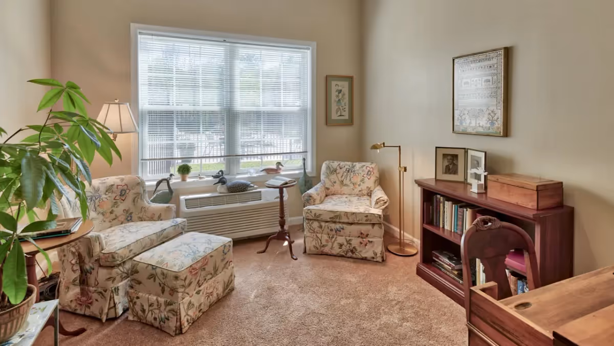 A cozy living room with two floral upholstered armchairs, one with a matching ottoman, positioned near a large window with blinds. A small wooden side table and a floor lamp are placed between the chairs. A wooden bookshelf with books, framed photos, and decorative items is against the wall. A large green potted plant is on the left side of the image.