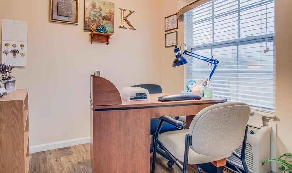A small office space with a wooden desk and two chairs, one on each side. On the desk are a blue adjustable desk lamp, a small glass container with pencils, and a white device. The room has light-colored walls with framed pictures and a large letter 'K' hanging on the wall. A window with white blinds lets in natural light, and a wooden floor is visible.