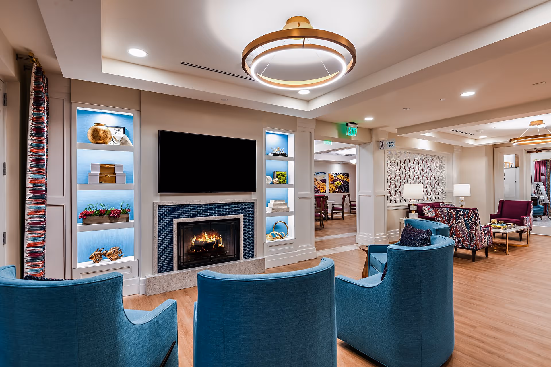 A modern and cozy living room area with a lit fireplace beneath a mounted flat-screen TV. The room features blue armchairs facing the fireplace, decorative shelves with various ornaments, and a stylish circular ceiling light. In the background, there is a dining room visible through an open doorway.