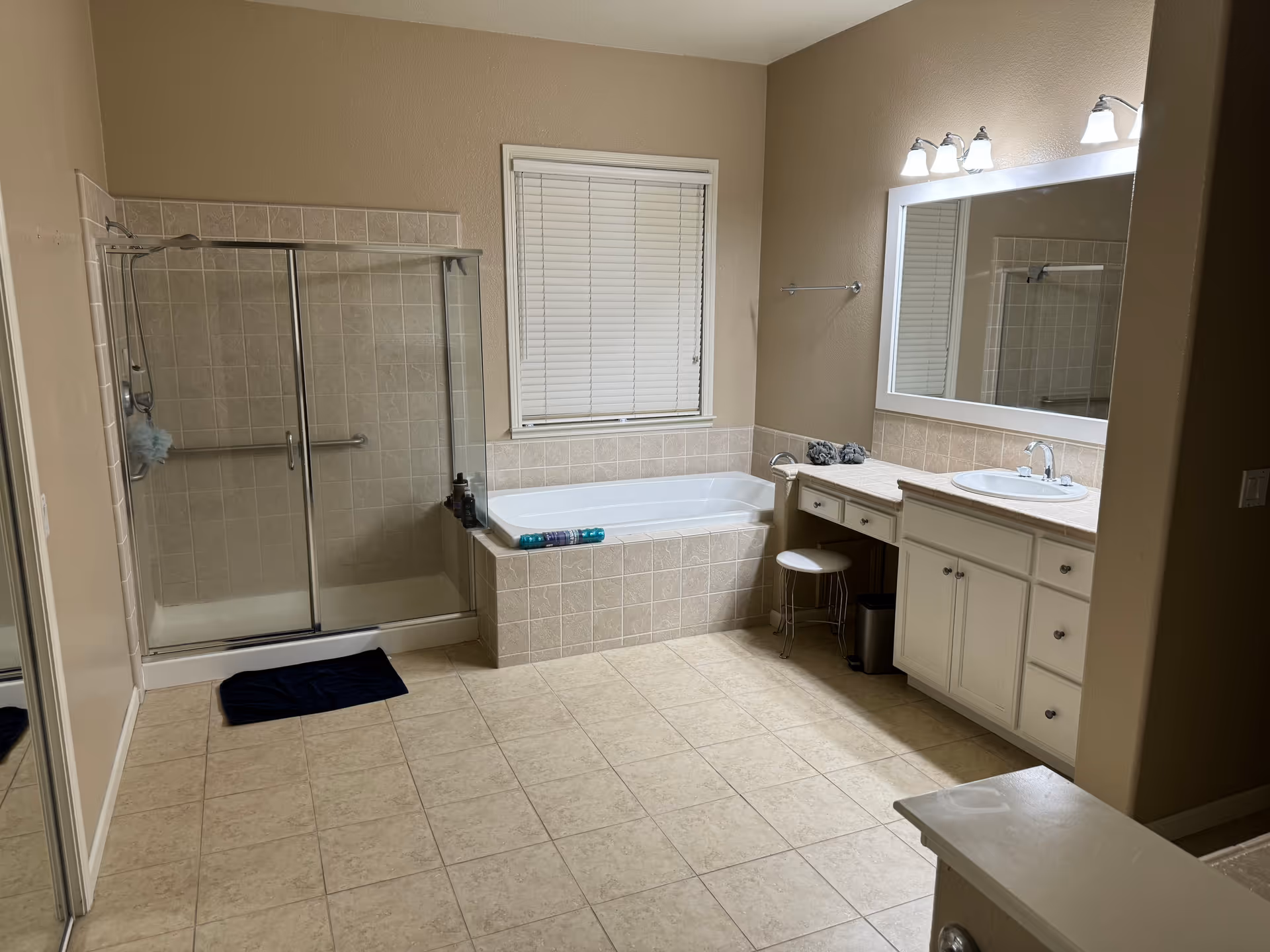 A spacious bathroom featuring a tiled floor, a glass-enclosed shower with a grab bar, a large bathtub with tiled surround beneath a window with closed blinds, and a vanity with a sink, mirror, and three light fixtures above. A small stool is tucked under the vanity, and a dark bath mat is placed in front of the shower.