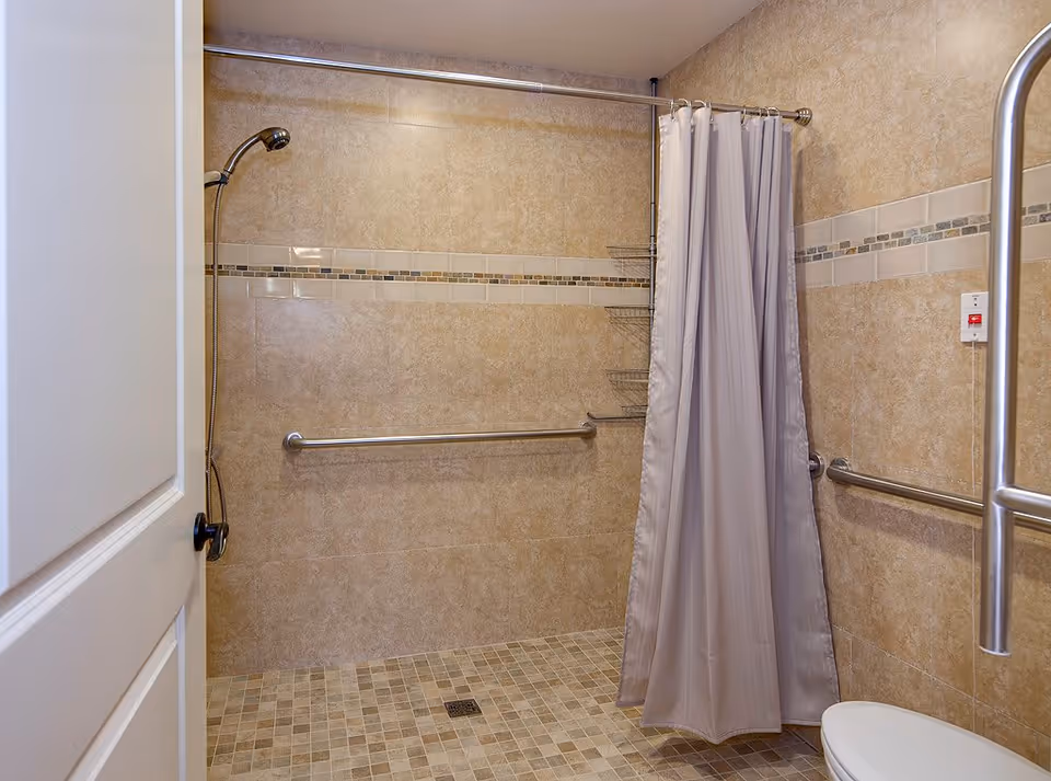 Accessible tiled shower with grab bars, handheld showerhead, shower curtain, and tiled floor drain.