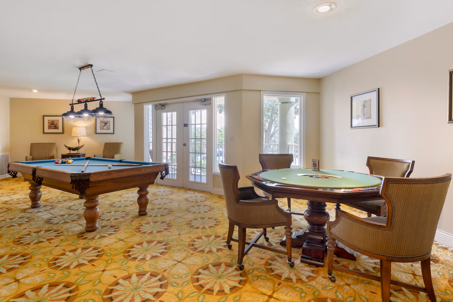 A recreational room with a pool table and a round poker table with four chairs. The room has patterned carpet, framed artwork on the walls, and a set of glass double doors leading outside. Two armchairs and a lamp are visible in the background near the pool table.