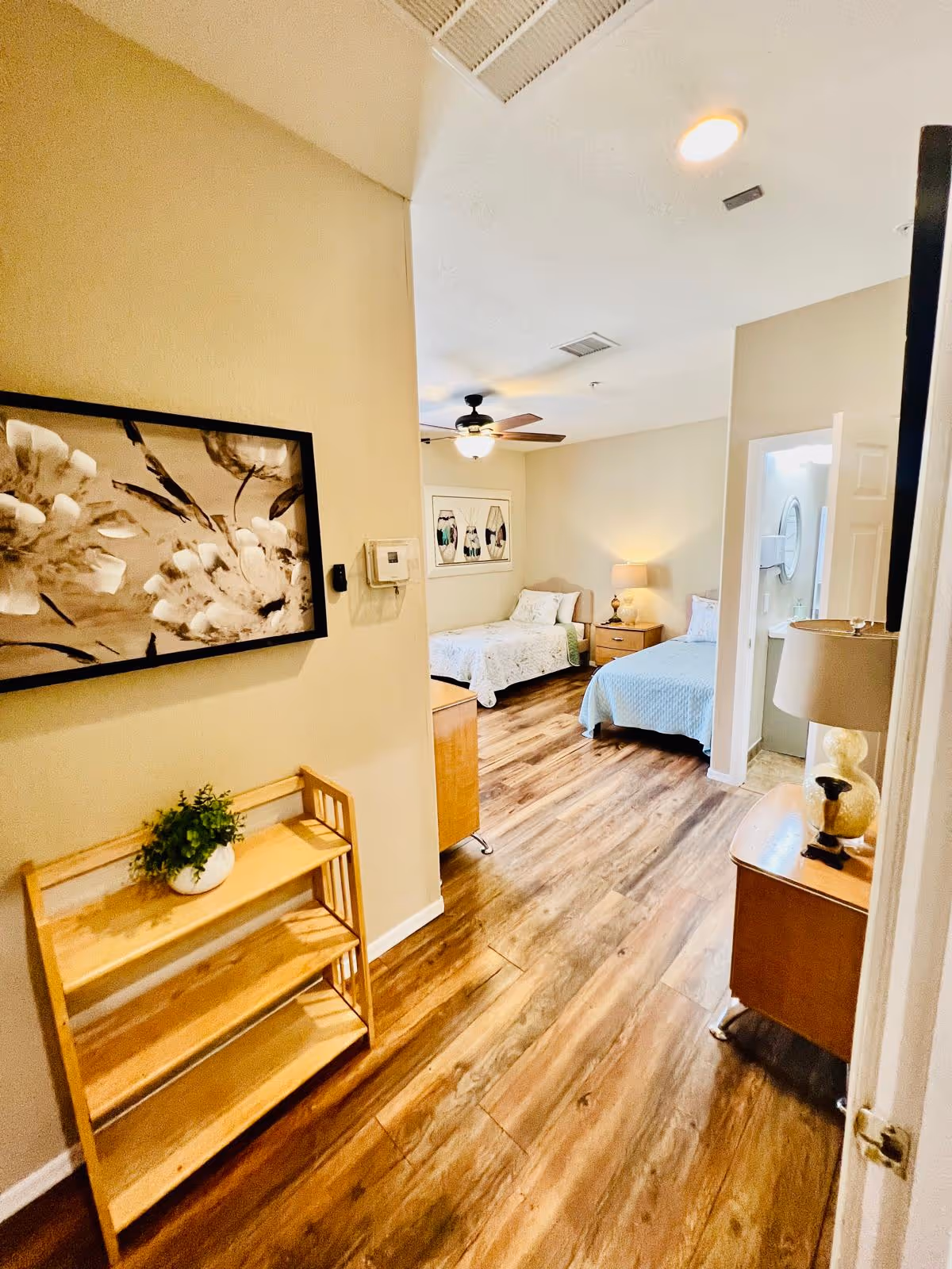 View down a hallway into a bedroom with two beds, each with a nightstand and lamp. The room has wood flooring, beige walls, a ceiling fan with light, and framed artwork on the wall. A small wooden shelf with a potted plant is visible in the foreground.