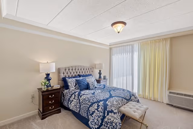 A well-lit bedroom with a large bed featuring a tufted headboard and blue and white patterned bedding. There are two dark wooden nightstands on either side of the bed, each with a lamp and decorative items. A small cushioned bench is placed at the foot of the bed. The room has beige walls, a carpeted floor, and a large window covered with sheer white and yellow curtains. An air conditioning unit is visible beneath the window.