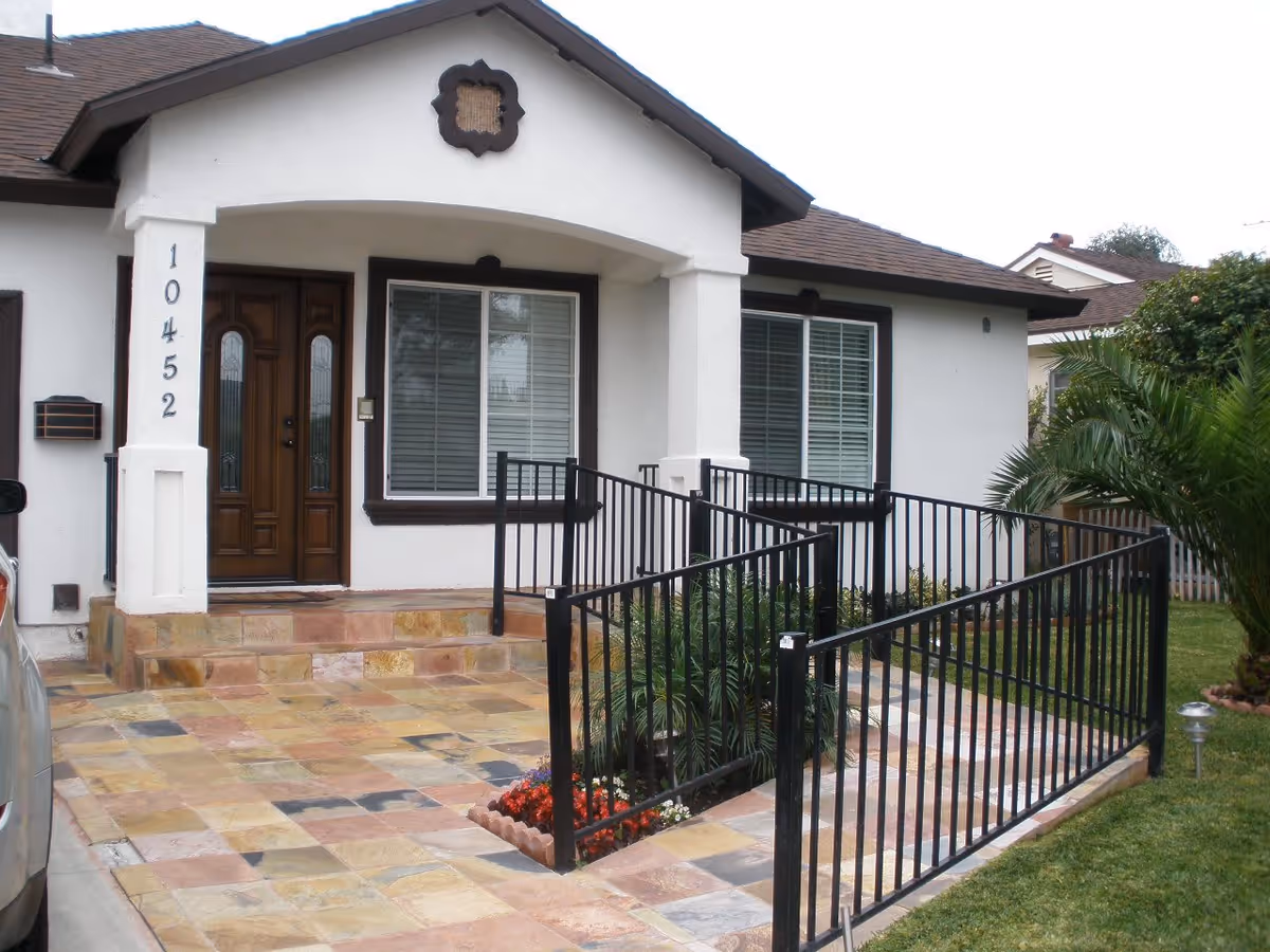 Front exterior view of a single-story building with white walls and brown trim. The entrance features a wooden door with glass panels, two windows with white blinds, and a tiled porch with a black metal railing. There is a small garden area with plants and flowers near the entrance.