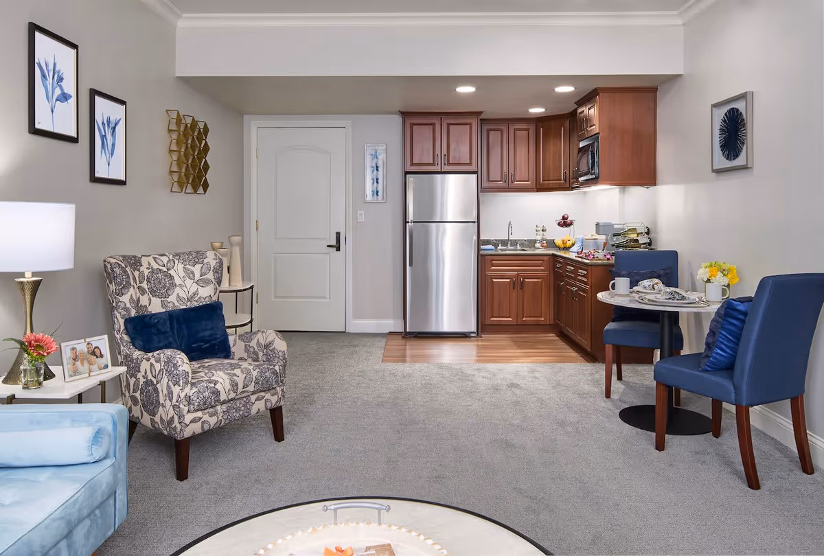 A cozy living space featuring a small kitchen with wooden cabinets and stainless steel refrigerator, a round dining table with two blue chairs, a floral patterned armchair with a blue cushion, a light blue sofa, and decorative wall art. The room has a mix of carpet and wood flooring and is softly lit with ceiling lights and a table lamp.