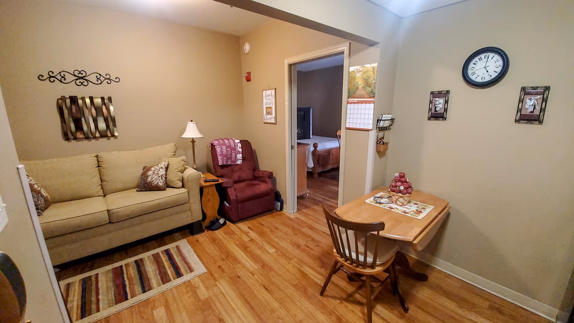A cozy living area in a senior living facility featuring a beige sofa with decorative pillows, a maroon recliner chair with a throw blanket, a small wooden side table with a lamp, and a wooden dining table with a single chair. The walls are decorated with framed pictures, a clock, and a metal wall art piece. The floor is wooden, and there is a doorway leading to a bedroom with a visible bed and TV.