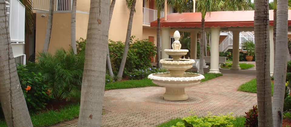 A landscaped courtyard featuring a three-tiered stone fountain, palm trees, and a covered colonnade beside the building.