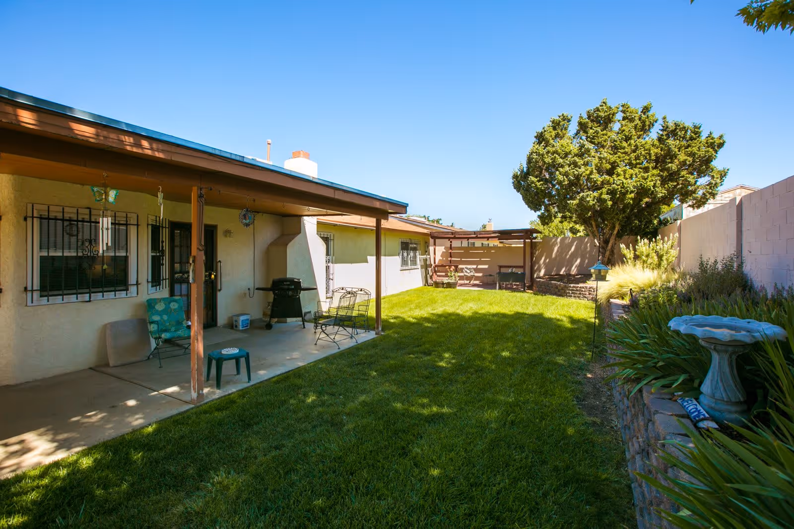 A sunny backyard area with a covered patio attached to a beige building. The patio has outdoor chairs, a small table, and a barbecue grill. The yard features a well-maintained green lawn, a large tree, and various plants along a stone-bordered garden bed. A birdbath is visible on the right side near the garden bed.