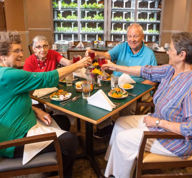 Four elderly residents seated around a dining table clinking glasses over salads in a bright dining room with shelves of plants in the background.