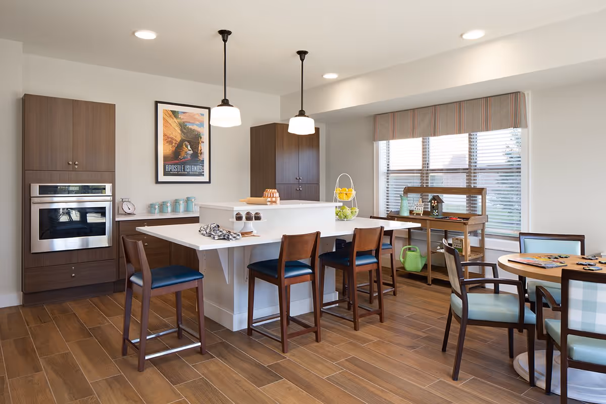 A modern kitchen and dining area with wooden flooring, a white island with four wooden chairs, two pendant lights hanging above the island, a built-in oven, cabinets, a round table with four chairs, and a large window with blinds letting in natural light. There are decorative items on the counter and shelves, including a tiered fruit basket, a watering can, and a small house model.