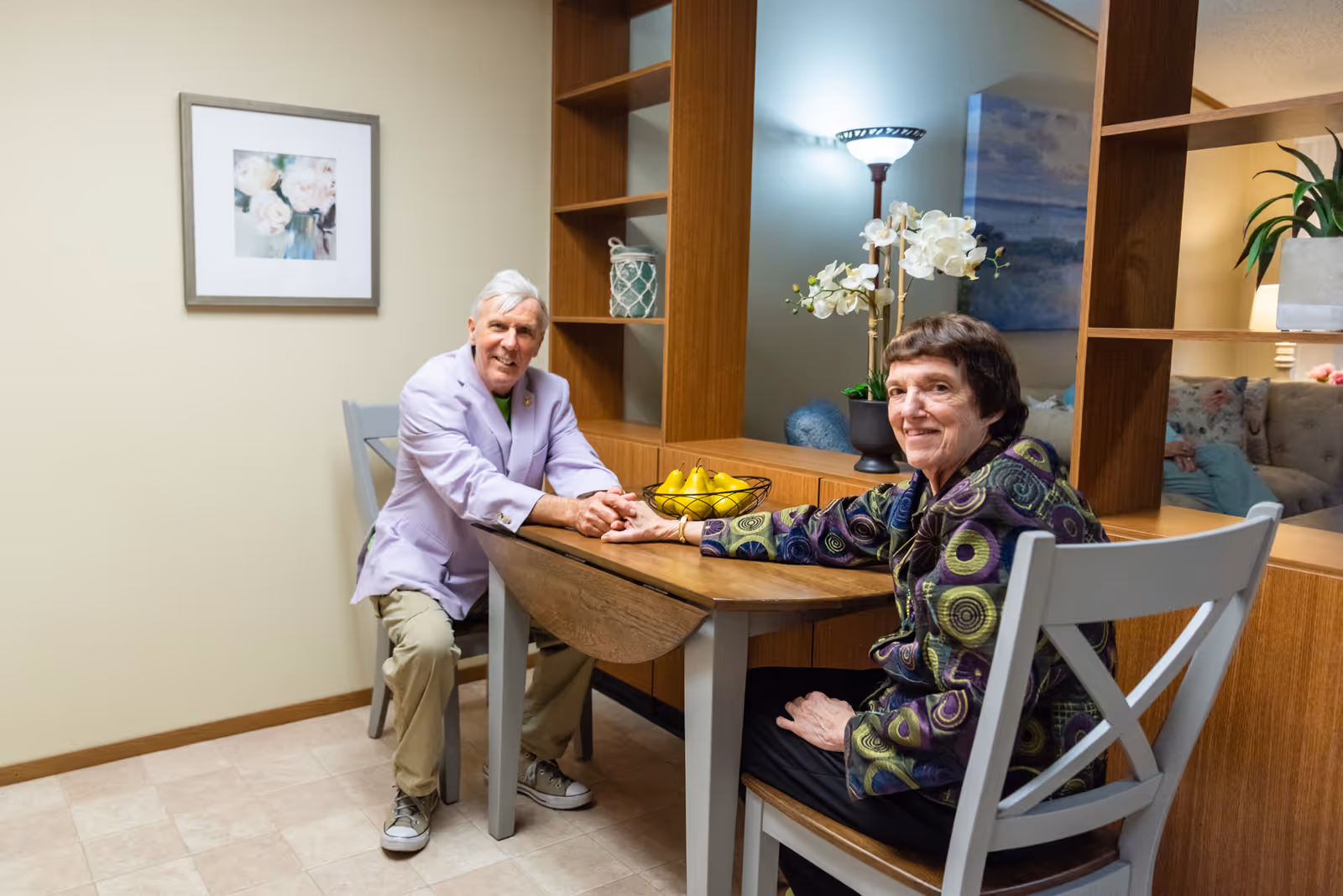 Two elderly people sitting at a wooden dining table in a cozy room. They are holding hands and smiling at the camera. The room has a wooden shelving unit with decorative items, a lamp, and framed artwork on the wall.