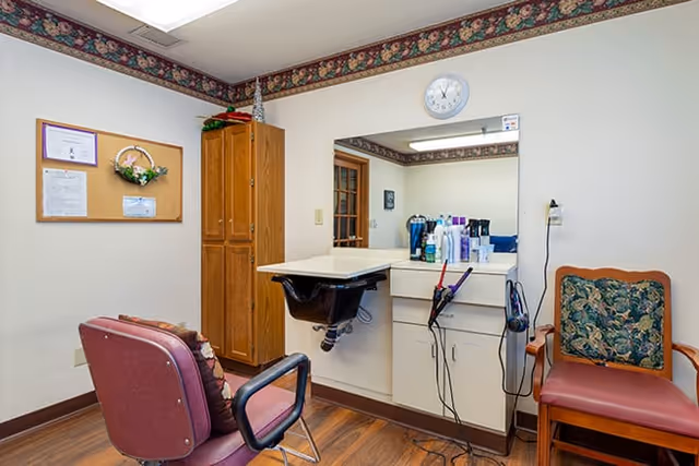 Interior view of a small salon or grooming area in a senior living facility with a salon chair, a wall-mounted mirror, a counter with various hair care products, a wooden cabinet, a bulletin board, and a chair with floral upholstery.