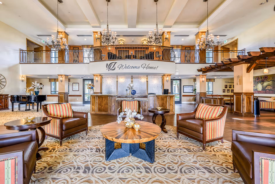 Spacious, elegantly furnished senior living lobby with seating, chandeliers, and a 'Welcome Home!' sign above the reception desk.