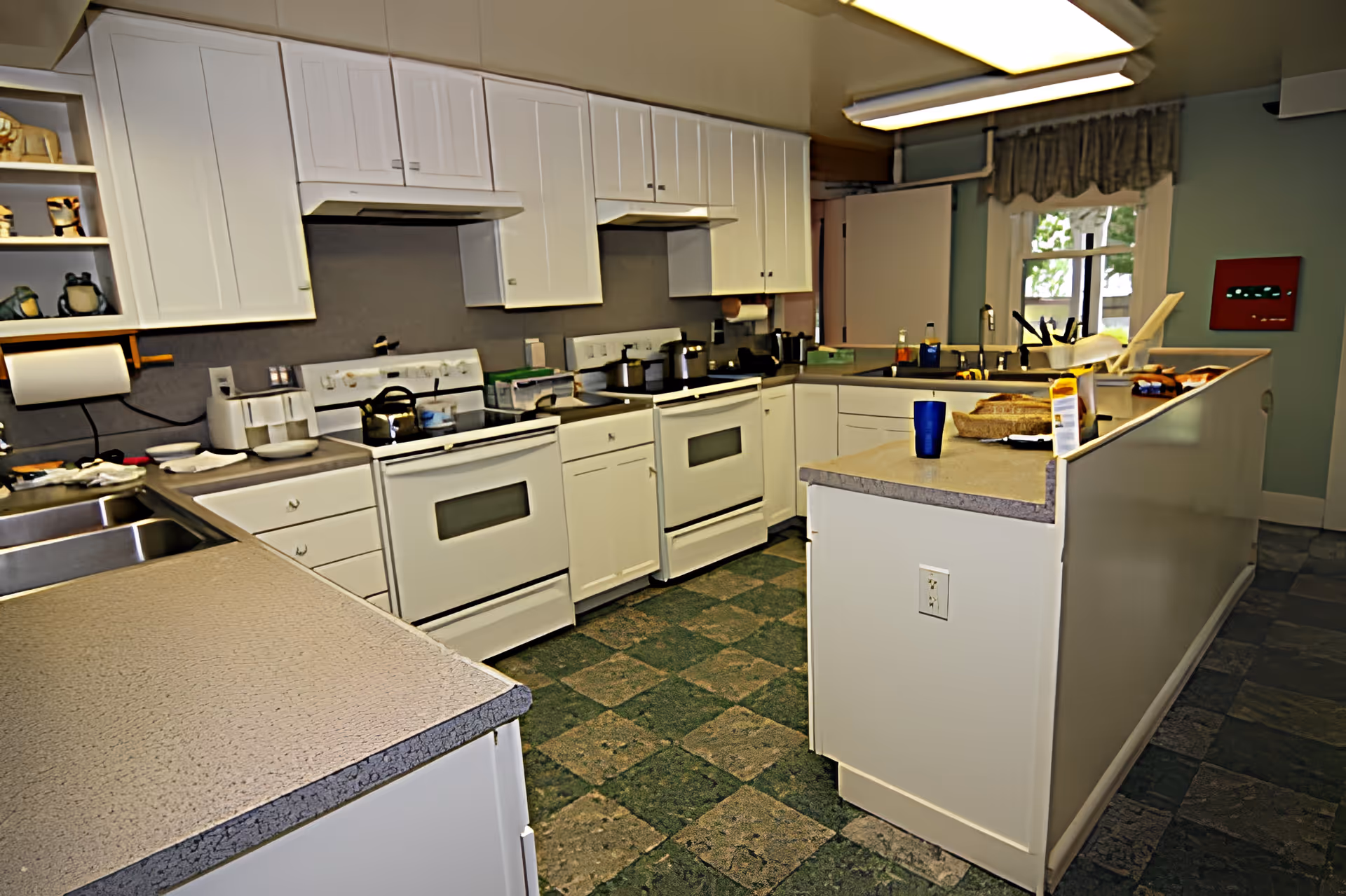 A spacious kitchen with white cabinets and countertops, featuring two white electric stoves, a double sink, various kitchen utensils, and a window with a valance. The floor has a green and gray checkered pattern, and there are overhead fluorescent lights.