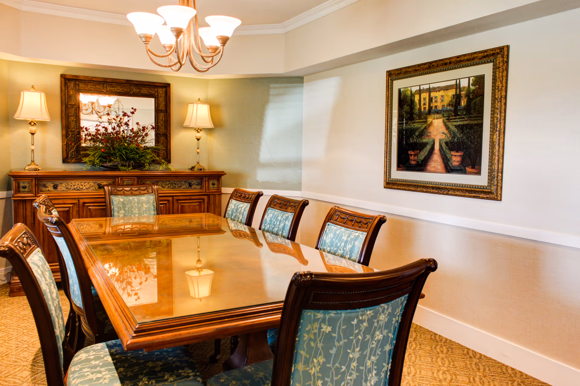 A formal dining room with a polished wooden table surrounded by eight upholstered chairs with blue floral fabric. The room features a wooden sideboard with two lamps and a large mirror above it. A framed painting hangs on the wall, and a chandelier with multiple lights is mounted on the ceiling.