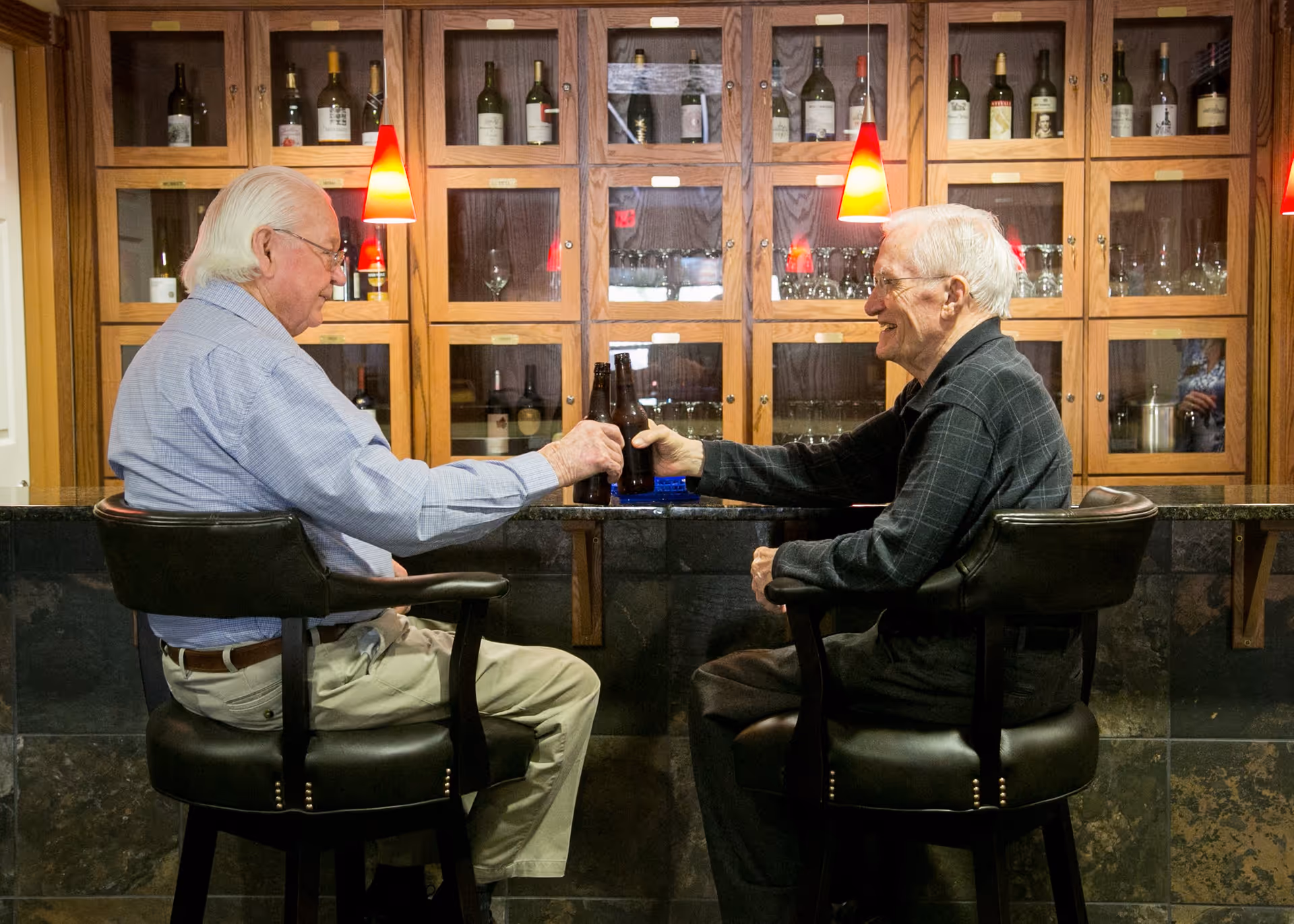 Two elderly men sitting on bar stools facing each other at a bar counter, clinking their beer bottles in a toast. Behind them is a wooden cabinet with glass doors displaying bottles and glassware, and red pendant lights hanging from the ceiling.