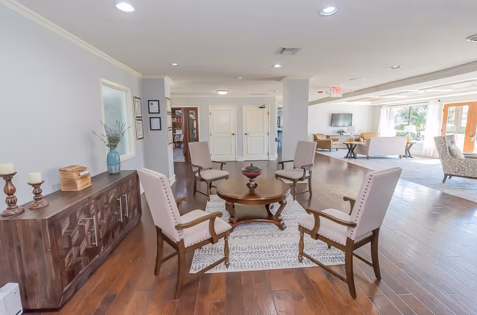 A bright and spacious senior living common area with four beige upholstered chairs arranged around a round wooden coffee table on a patterned rug. The room features wooden flooring, a wooden sideboard with decorative items including candles and a vase with flowers, and a large window letting in natural light. In the background, there are additional seating areas with sofas and armchairs, a wall-mounted TV, and glass doors leading outside.