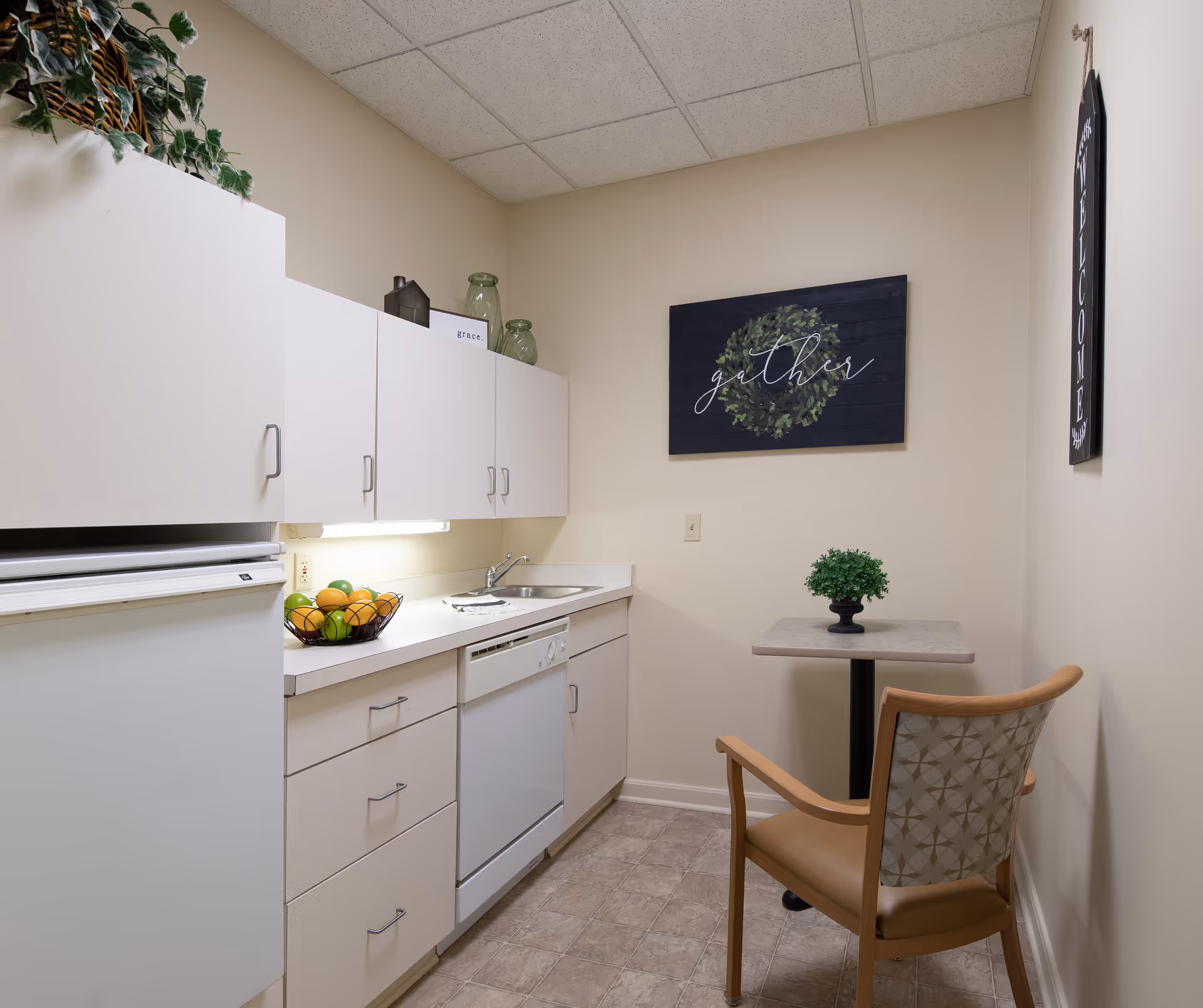 A compact kitchenette with white cabinets, a sink and dishwasher, a fruit bowl on the counter, and a small table with a chair under wall decor.