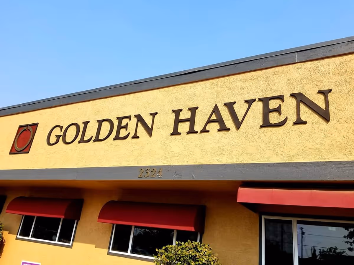 Exterior view of a building with a yellow textured wall and a sign that reads 'GOLDEN HAVEN' in large brown letters. The building has red awnings above the windows and the number 2324 is displayed below the sign. The sky is clear and blue.