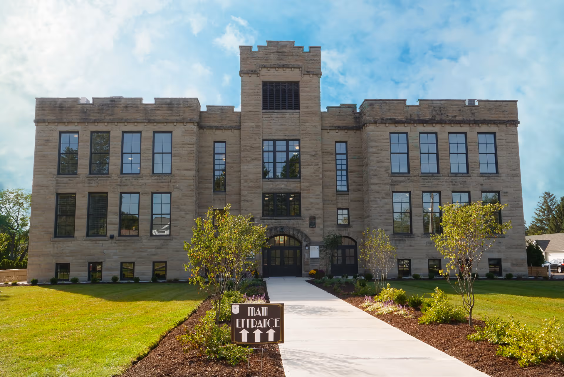 Stone three-story historic school building with a central tower, landscaped lawn, walkway, and a 'Main Entrance' sign.