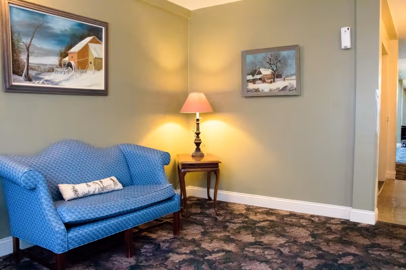 A cozy corner of a room with a blue upholstered loveseat, a small wooden side table with a lamp emitting warm light, and two framed winter landscape paintings on light green walls. The floor is covered with a patterned carpet.