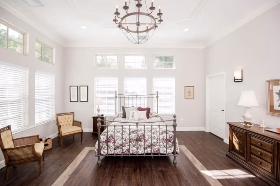 A bright and spacious bedroom with a metal bed frame and floral bedding centered against a wall with multiple windows. The room features dark wood flooring, two beige upholstered chairs on the left, a wooden dresser with a lamp and framed artwork on the right, and a chandelier hanging from the ceiling.