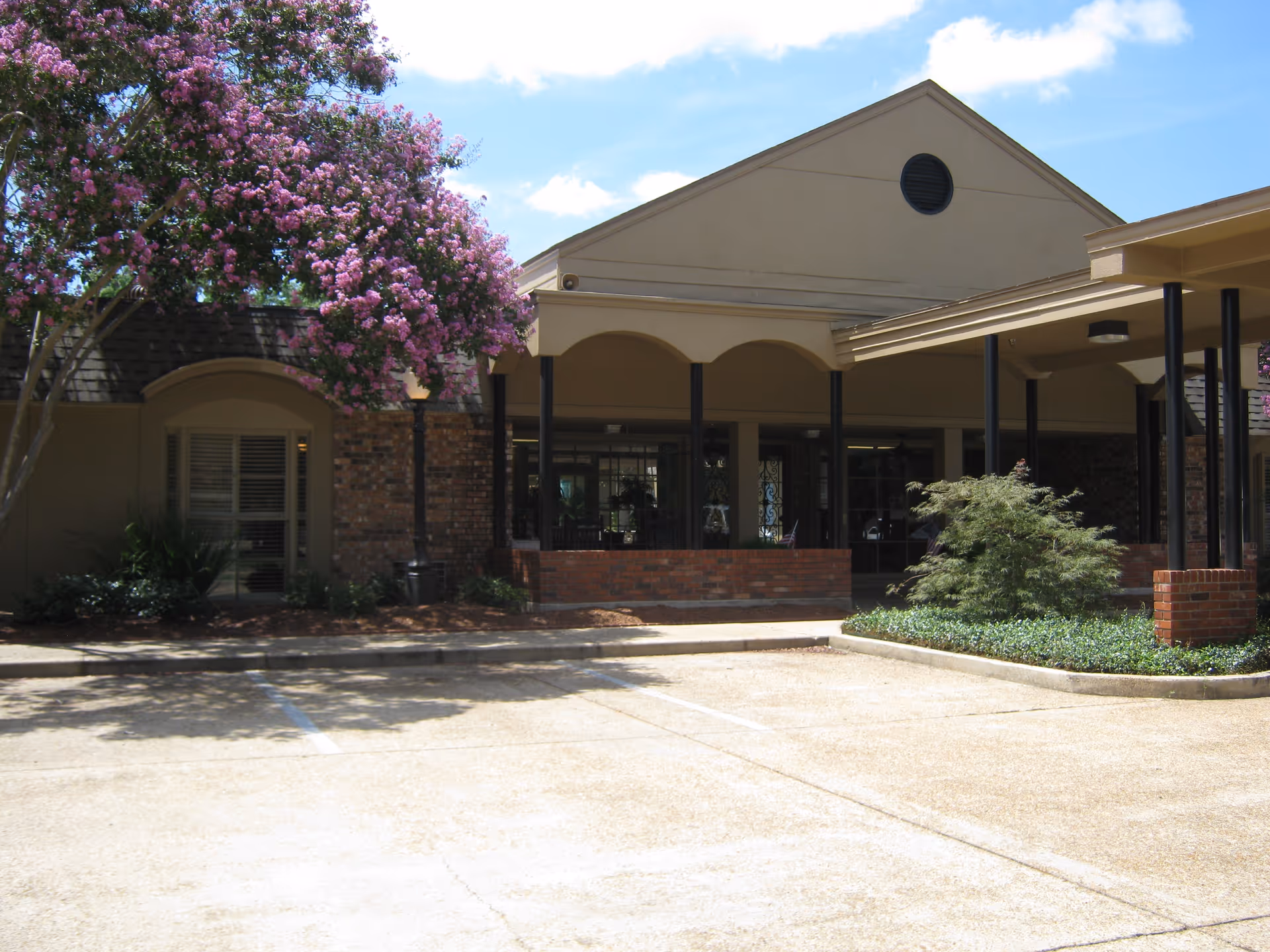 Exterior view of Matthews Memorial Health Care building with a covered entrance, brick walls, and a flowering tree with pink blossoms on the left side. The sky is partly cloudy and there is a paved parking area in front.