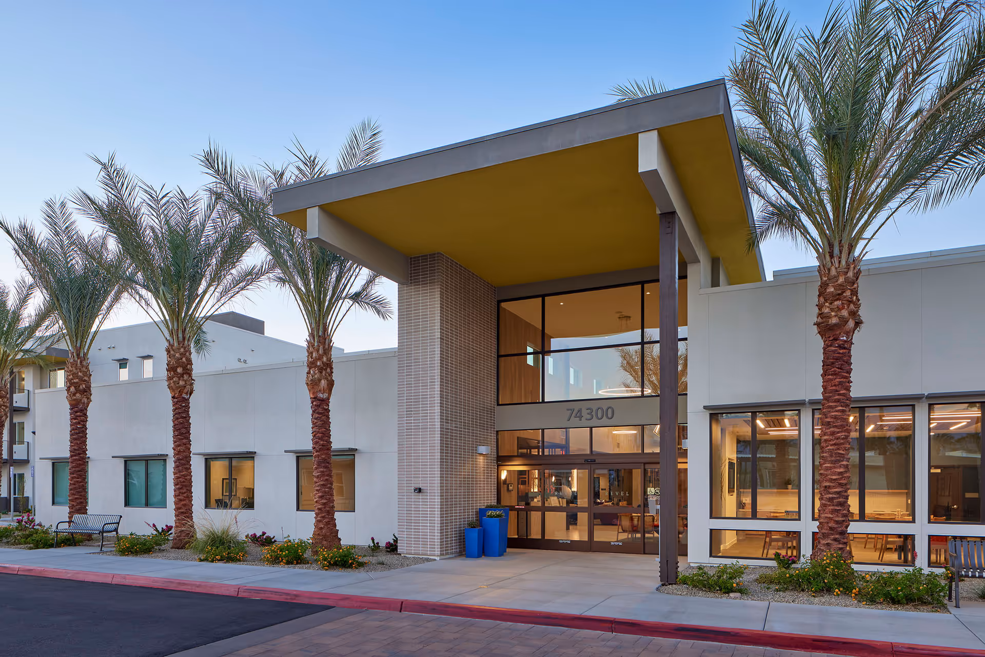 Modern senior living facility entrance with a large yellow-overhang canopy, glass double doors marked 74300, and palm trees lining the forecourt.