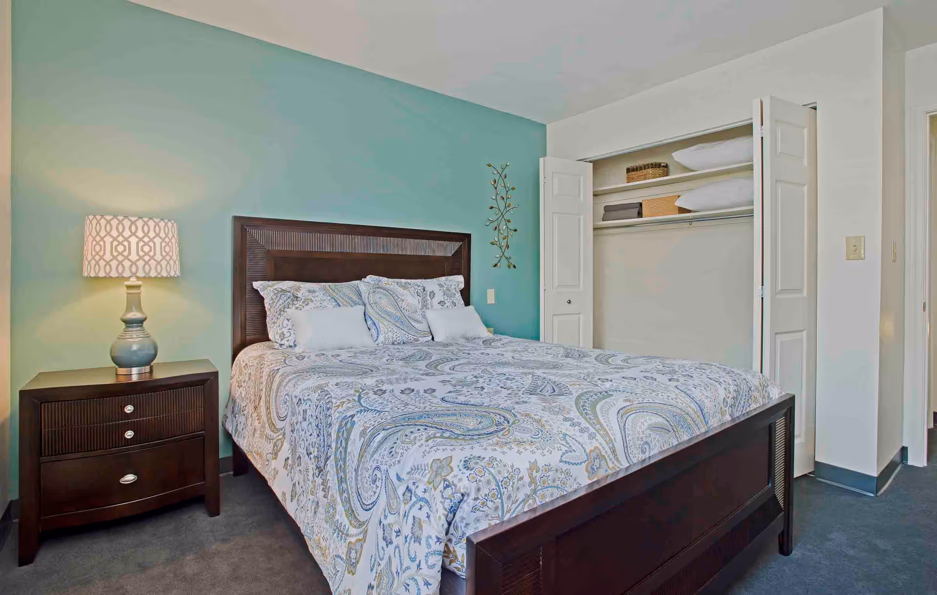 A bedroom with a dark wooden bed frame and a paisley-patterned bedspread in shades of blue, white, and beige. There is a matching dark wooden nightstand with a decorative lamp on it to the left of the bed. The wall behind the bed is painted teal, and there is a metal wall decoration. To the right, there is an open closet with shelves holding pillows and baskets.
