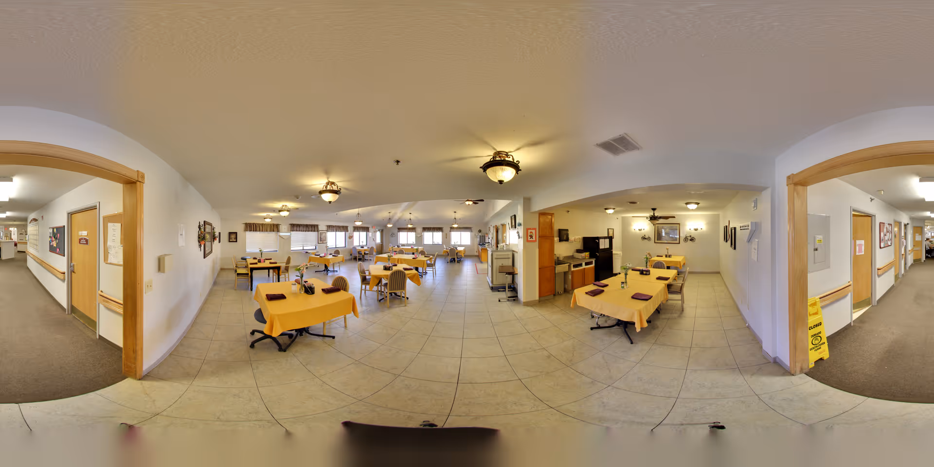 Large dining room with multiple tables covered in yellow tablecloths, chairs, and a serving/kitchen area in a senior living facility.