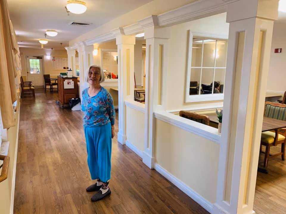 An elderly woman with short white hair wearing a blue floral top and blue pants stands smiling in a well-lit hallway with wooden flooring. The hallway has cream-colored walls with decorative columns and windows looking into a dining area with tables and chairs. There are ceiling lights and chairs along the hallway.