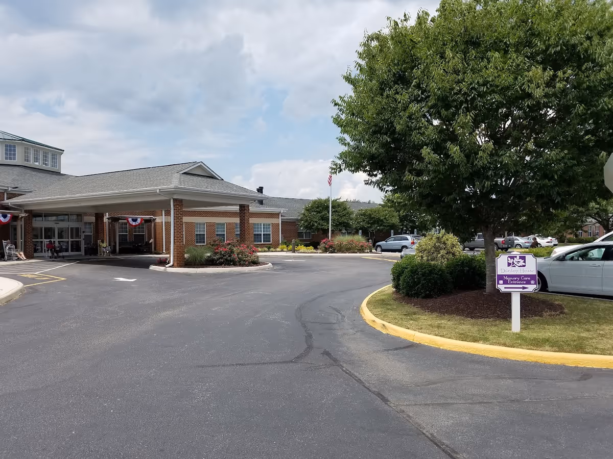 Exterior view of Dunlop House Assisted Living & Memory Care building with a covered entrance, a driveway, landscaped bushes and trees, parked cars, and a sign indicating the Memory Care Entrance.
