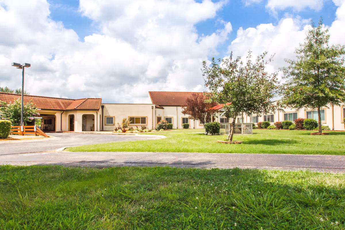 Exterior view of a single-story building with a red-tiled roof, surrounded by green grass, trees, and a paved driveway under a partly cloudy sky.