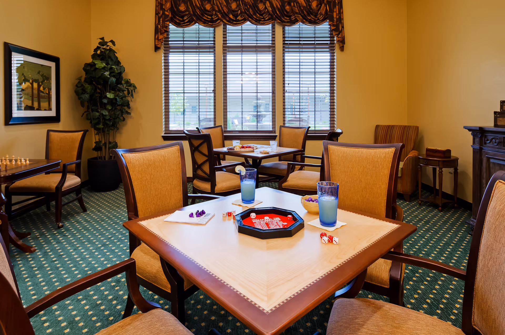 A cozy game room with several wooden tables and cushioned chairs. On the nearest table, there are dice, game pieces, and two glasses of a light blue beverage with lemon slices. The room has green carpet with a dotted pattern, beige walls, a large window with blinds and a valance, a potted plant, framed artwork, and a striped armchair in the corner.
