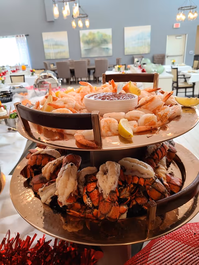 A two-tiered serving tray with cooked shrimp and lobster tails, garnished with lemon wedges and a bowl of cocktail sauce on the top tier. The background shows a dining area with tables, chairs, and wall art in a senior living community.