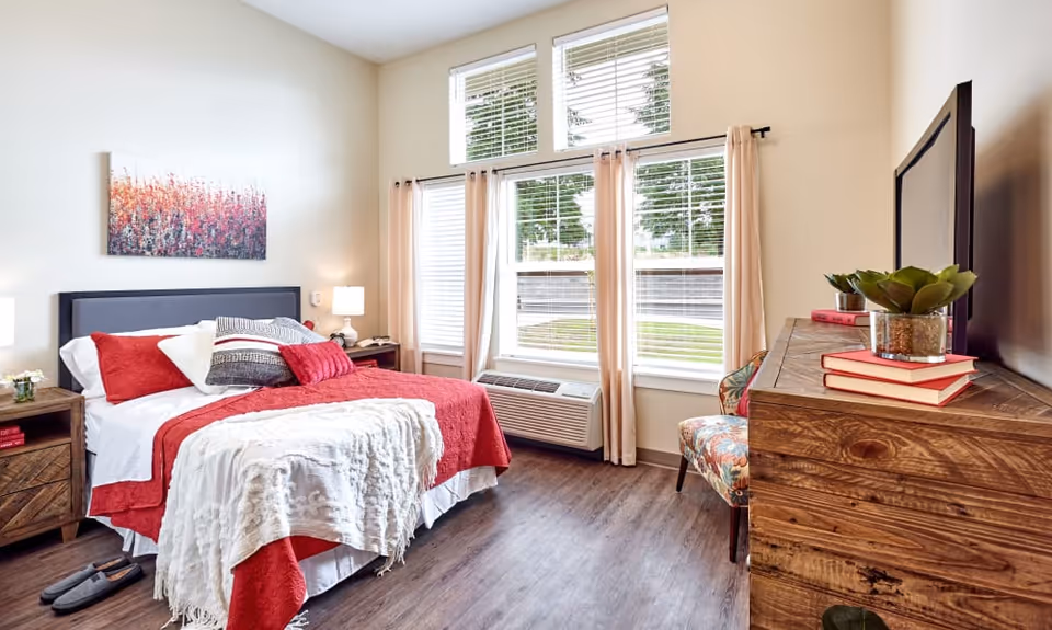 A bright and cozy bedroom with a bed dressed in red and white linens, multiple pillows, and a white throw blanket. There are two wooden nightstands with lamps on either side of the bed. A colorful floral-patterned chair is placed near a large window with beige curtains and white blinds. A wooden dresser with a flat-screen TV and decorative items, including books and a small plant, is positioned opposite the bed. The room has wood flooring and light-colored walls with a piece of abstract art hanging above the bed.