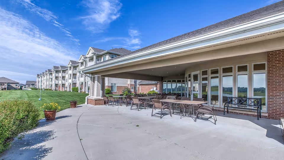 Covered outdoor patio with tables and chairs beside a multi-story assisted living building and a grassy lawn under a blue sky.