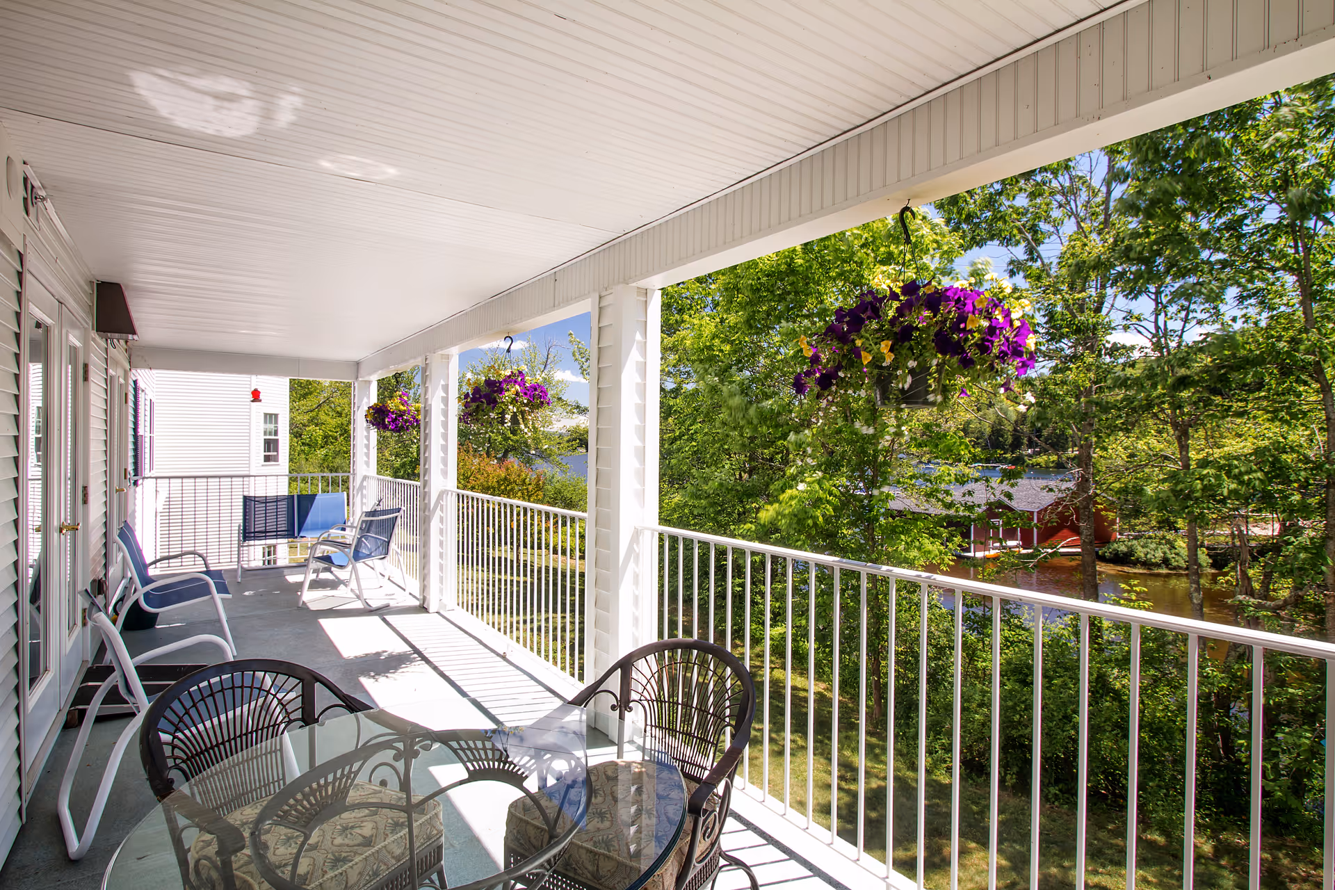 A covered outdoor balcony with white railings and ceiling, featuring several chairs and a glass-top table. Hanging baskets with purple flowers are suspended from the ceiling. The balcony overlooks a green, tree-filled landscape with a glimpse of a red building and a body of water in the background.