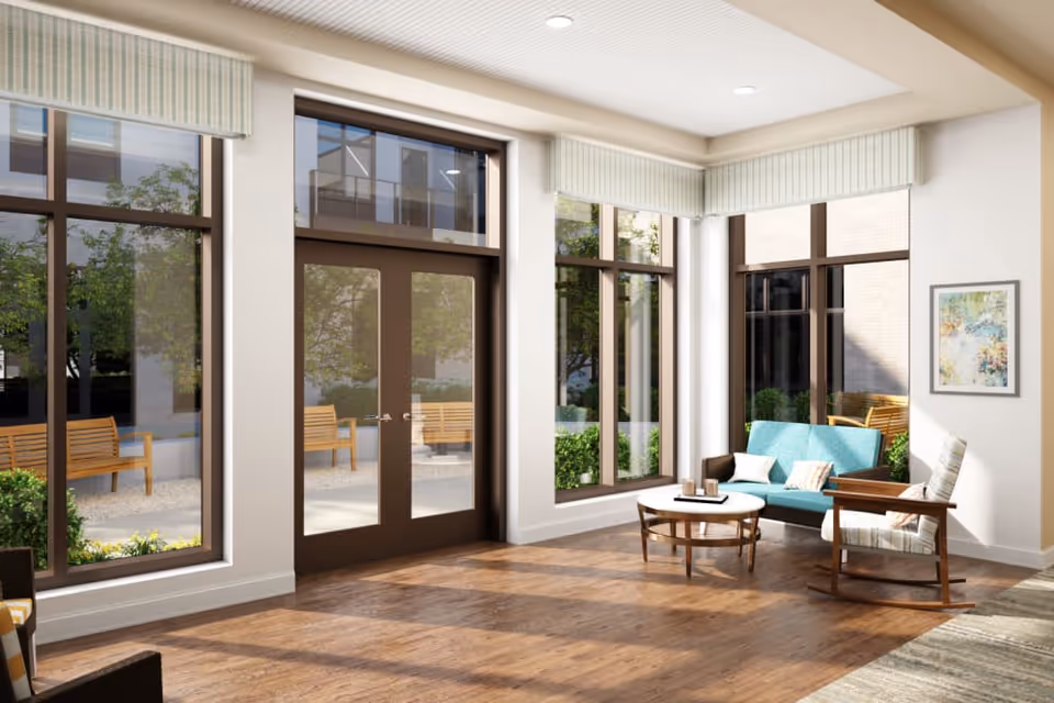 Bright lounge with large windows and glass doors opening to an outdoor bench area, featuring a teal sofa, wooden rocking chair, and round coffee table on hardwood floors.