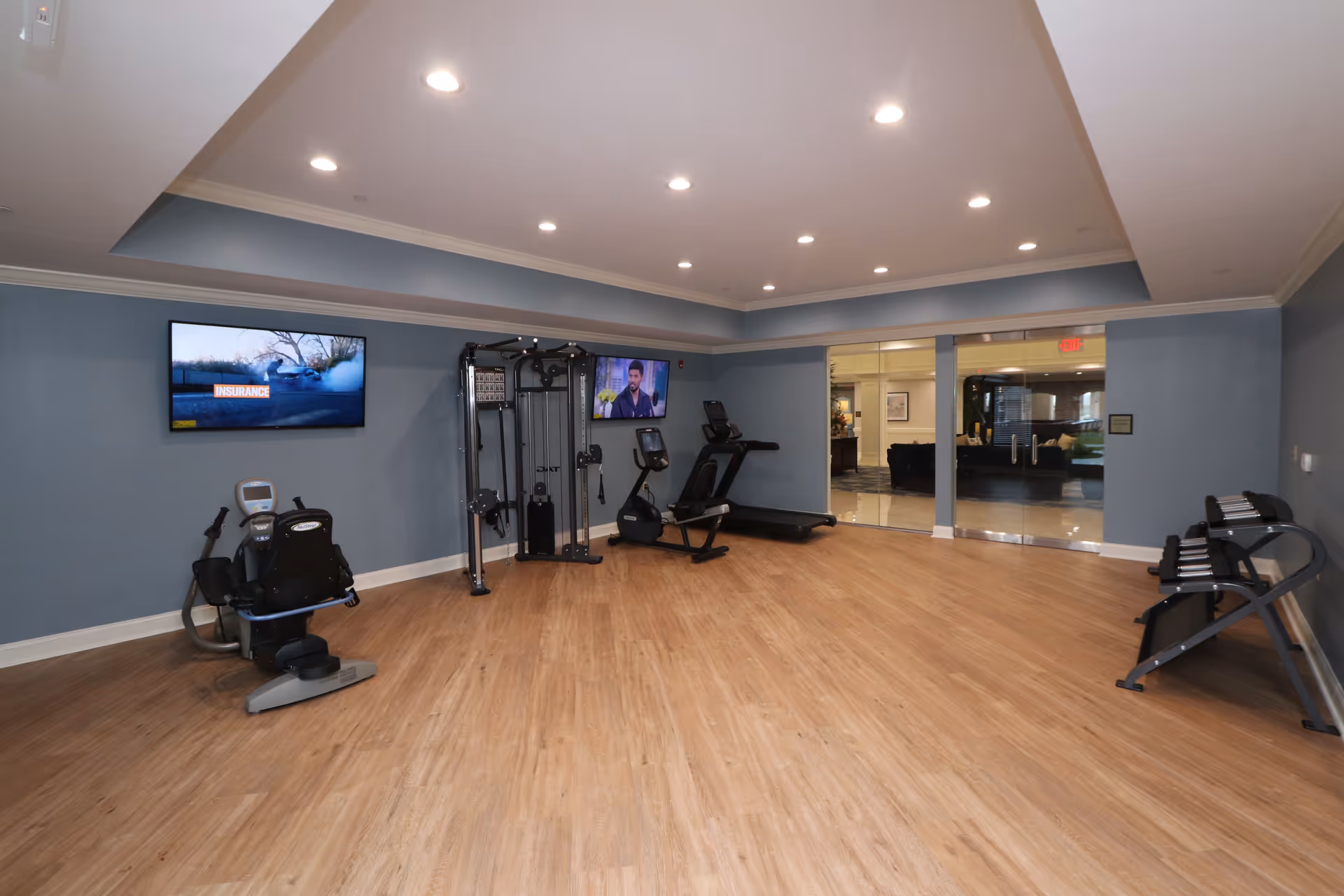 A fitness room with light wood flooring and blue-gray walls. The room contains exercise equipment including a recumbent bike, a multi-function weight machine, a treadmill, and a set of dumbbells on a rack. Two flat-screen TVs are mounted on the walls. The room is well-lit with recessed ceiling lights and has glass double doors leading to another area.