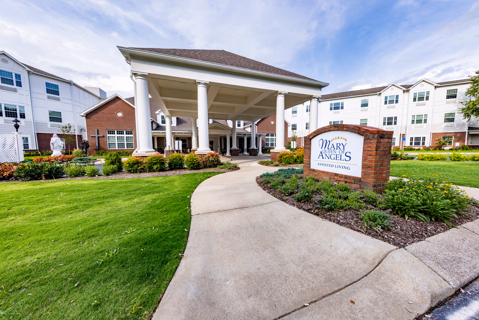 Exterior view of Mary Queen of Angels Assisted Living facility showing a covered entrance with white columns, a brick sign with the facility name, well-maintained green lawns, and a multi-story building in the background under a partly cloudy sky.